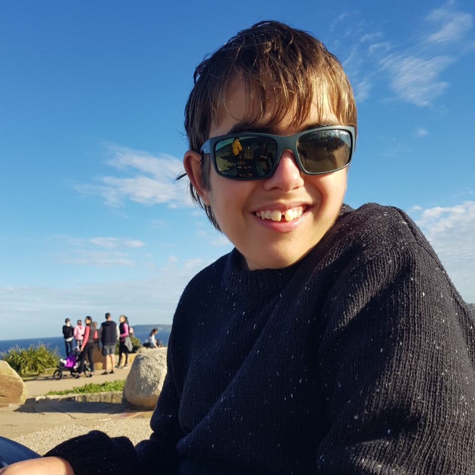 Photo of Luca Weber wearing sunglasses and smiling, a bright blue sky and coastline behind him.