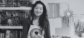 Portrait photo of Mai, sitting on a stool with glass rods in a shelf on her left and glass beads on a desk to her right