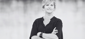 Photo of Suzie Dureau, standing in front of a blurred artwork, arms folded and smiling.