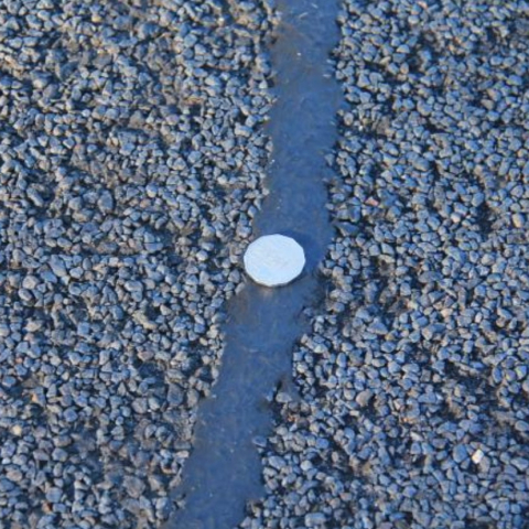 Close-up of an asphalt road surface with a sealed crack running vertically through the center. The crack is filled with dark sealing material, and a coin is placed on the sealed crack for scale, showing the size and texture of the sealant compared to the surrounding road.