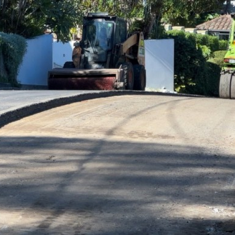 Road repair scene on a residential street with two pieces of heavy machinery. On the left, a compact track loader with a large brush attachment clears debris, while on the right, a vibratory roller operated by two workers compacts freshly laid asphalt. The foreground shows a partially milled road surface with exposed dirt and gravel. Lush greenery and residential houses are visible in the background.