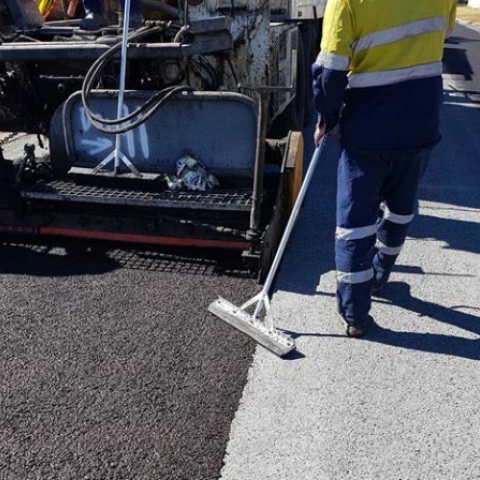 A worker in high-visibility clothing uses a long-handled tool to smooth a freshly laid layer of microsurfacing material on a road. The image shows a clear contrast between the dark, untreated road surface and the lighter, newly applied microsurfacing. Machinery for applying the microsurfacing is visible in the background.